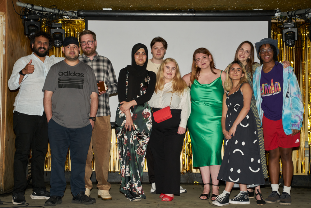 Comedian Nish Kumar, Arts Emergency founder Neil Griffiths and the Youth Collective which consists of 8 people aged between 18 and 28, standing on a stage. Photo taken at the 10th Birthday celebration at the MOTH Club, London. June 2023. Photo by Vanessa Ng