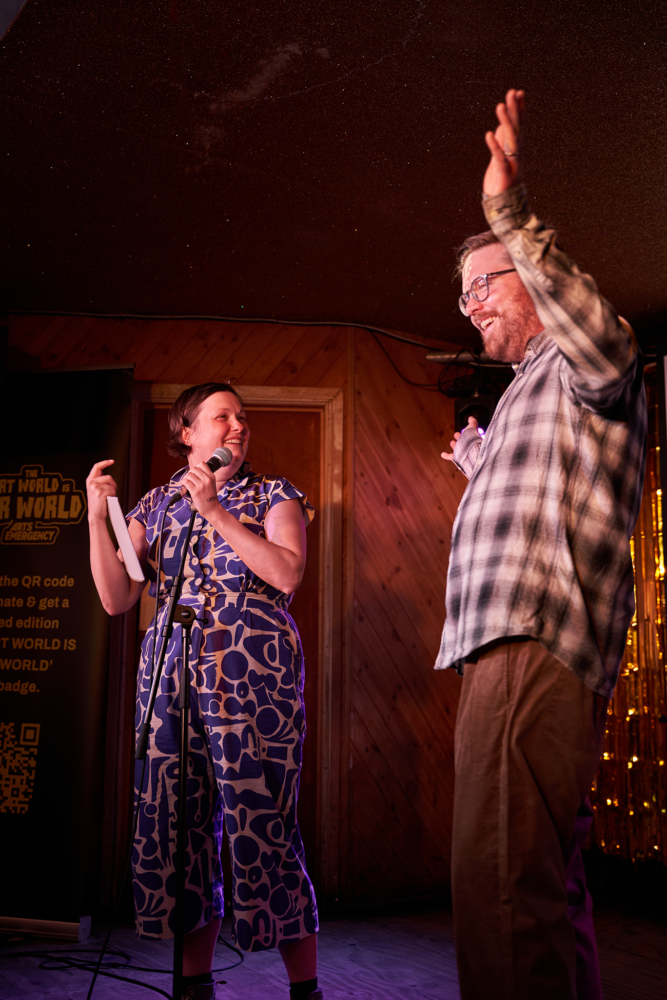 Comedian Josie Long stand on stage holding a microphone in a blue and white patterned jumpsuit looking at Neil Griffiths who is wearing a plaid shirt and tan slacks and throwing his arms up cheerfully. They're both grinning. Photo taken at the MOTH Club, London. June 2023. Photo by Vanessa Ng.