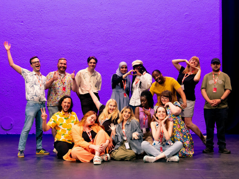 14 people, some standing some sitting, on a stage with a purple background. They are a variety of ages between 18 and 45 and look happy.