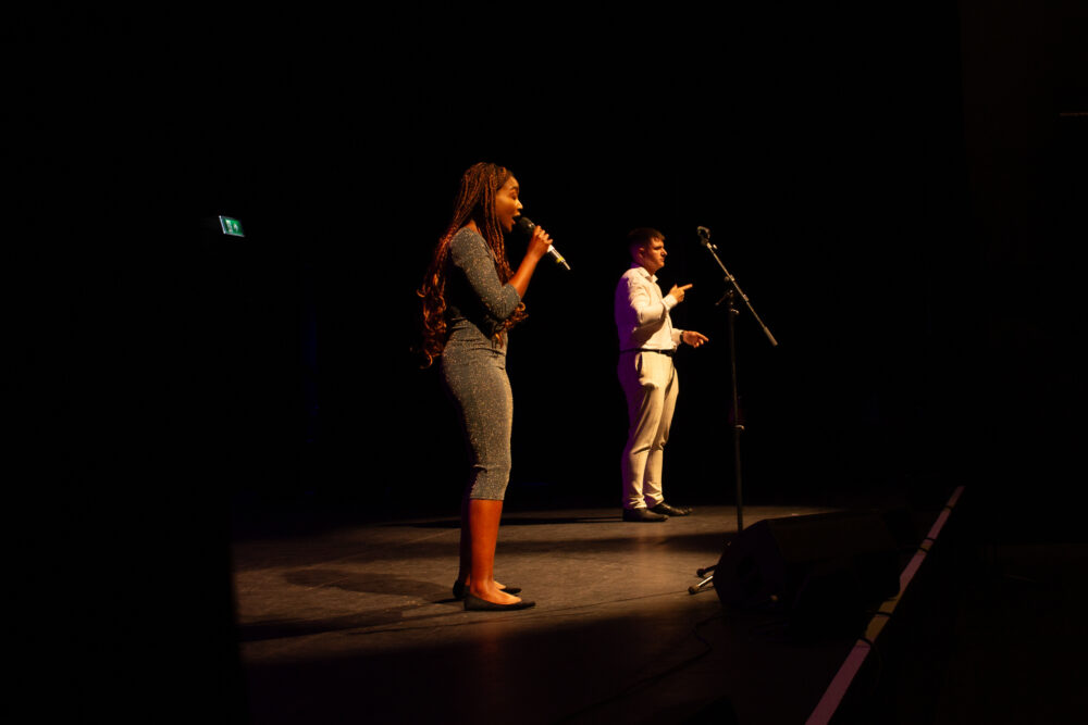 Singer B!TEZ standing on a theatre stage with a microphone singing. They are wearing a grey fitted dress. Photo taken at Youth Collective's The Art World Is Our World Festival at Contact, Manchester, June 2023. Photo by Ronke Hart