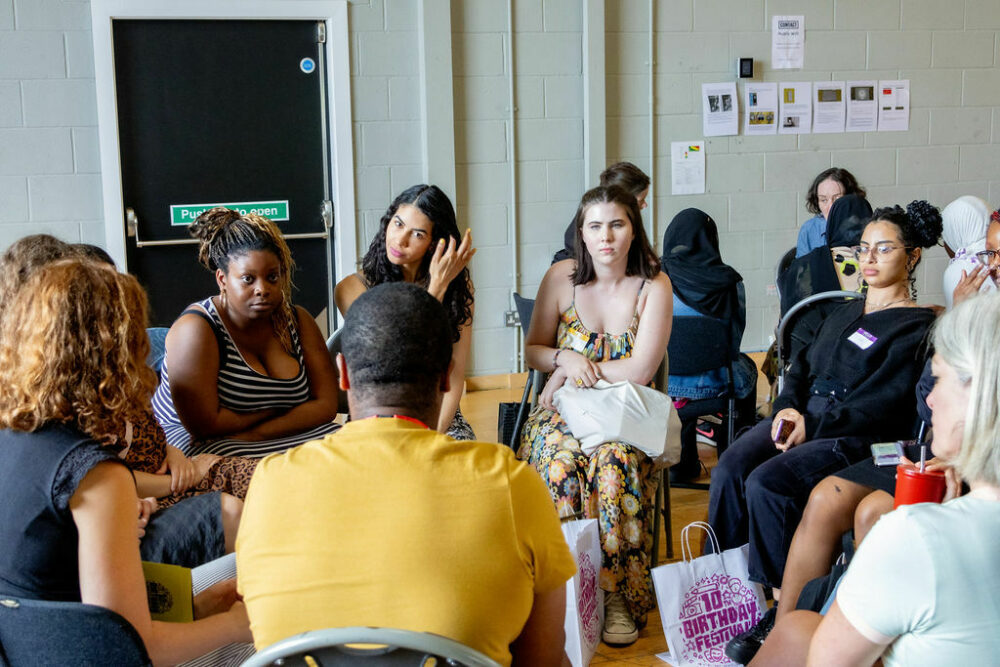 8 people sat in a circle of varying ages sat in a circle talking to each other. Photo taken at Youth Collective's The Art World Is Our World Festival at Contact, Manchester, June 2023. Photo by Ronke Hart