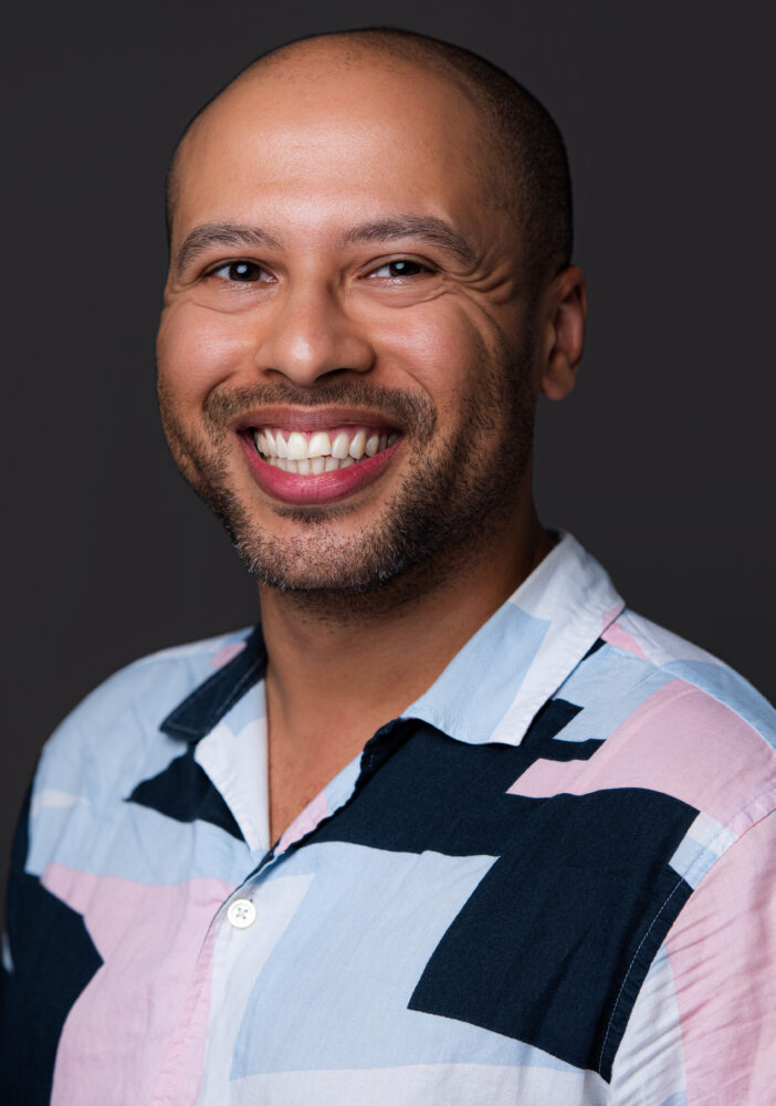 Headshot of Dean Atta - a mixed race male, smiling wearing a shirt with pastel coloured shapes on