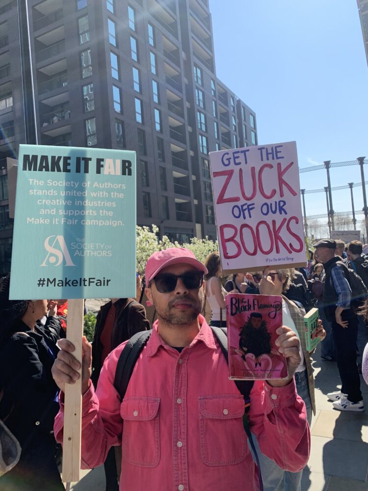 Dean Atta - mixed race man wearing a pink cap and jacket holding up placards in a protest against Meta AI