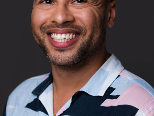 Headshot of Dean Atta - a mixed race male, smiling wearing a shirt with pastel coloured shapes on