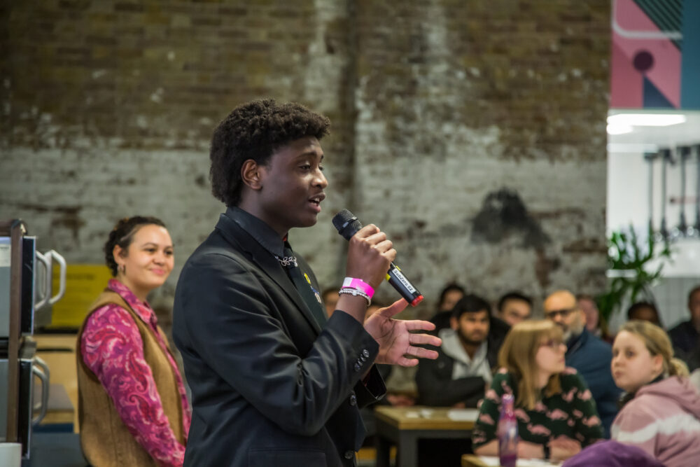 A young person wearing a smart black suit talking to a room of people of varying backgrounds and ages