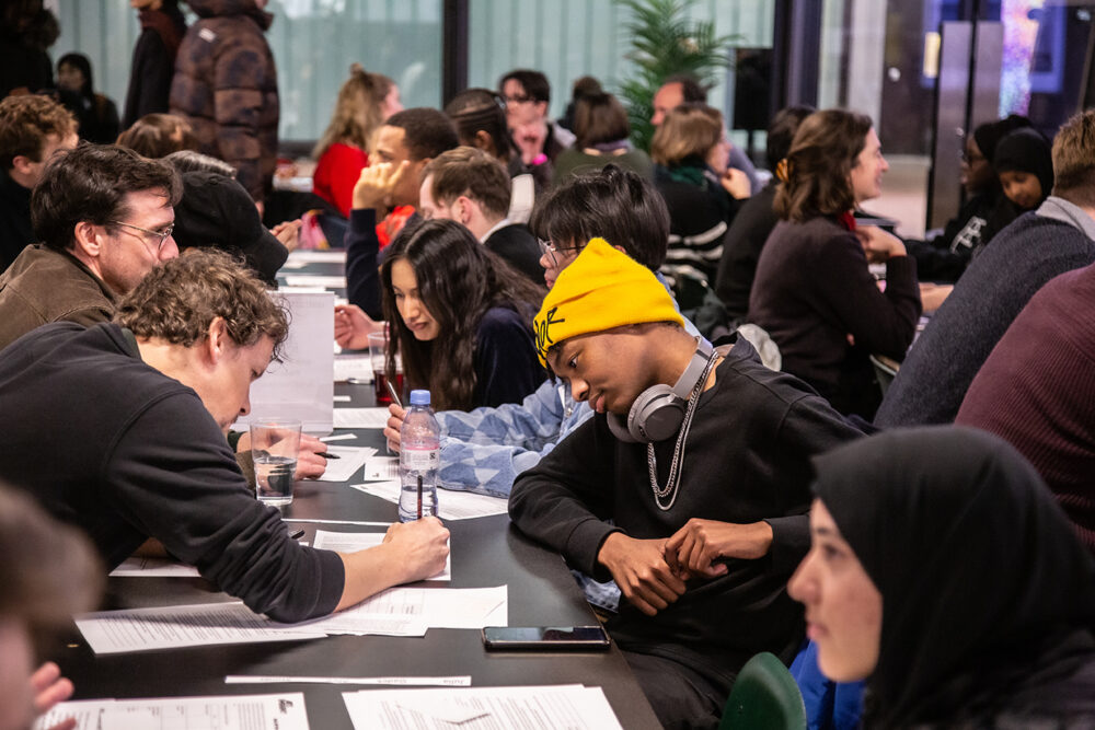A room full of people of varying ages and backgrounds at tables chatting and taking notes
