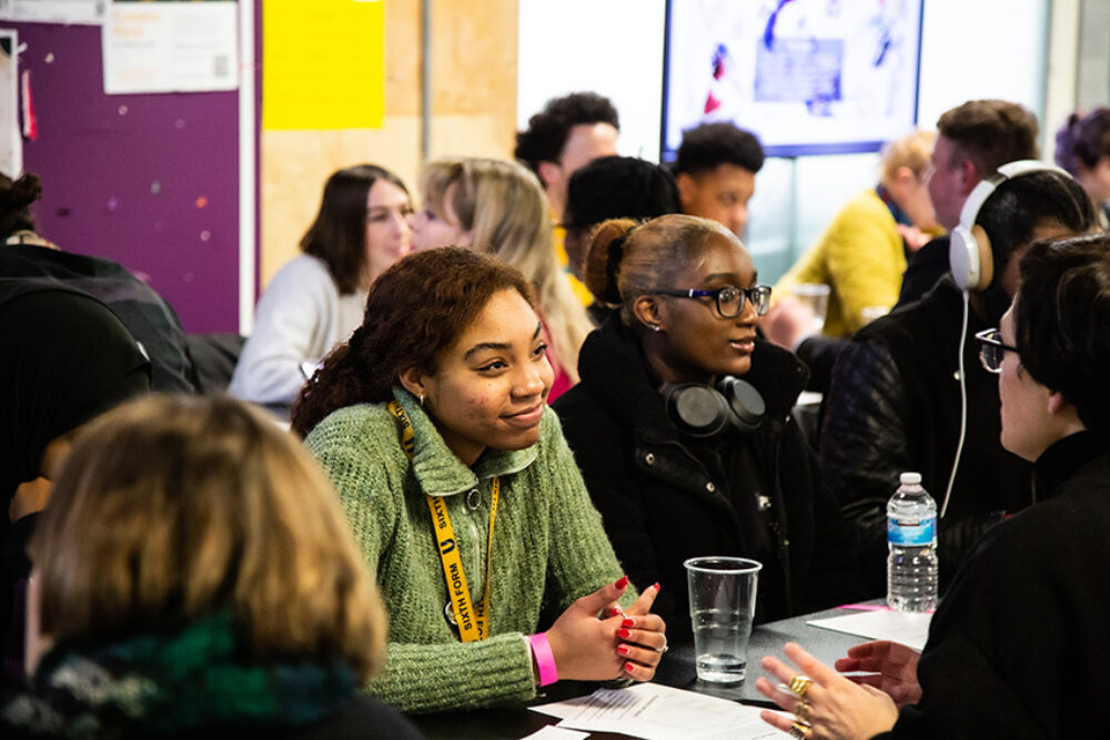 A young person with brown hair and wearing a green jumper looks engaged while listening to someone talking the them