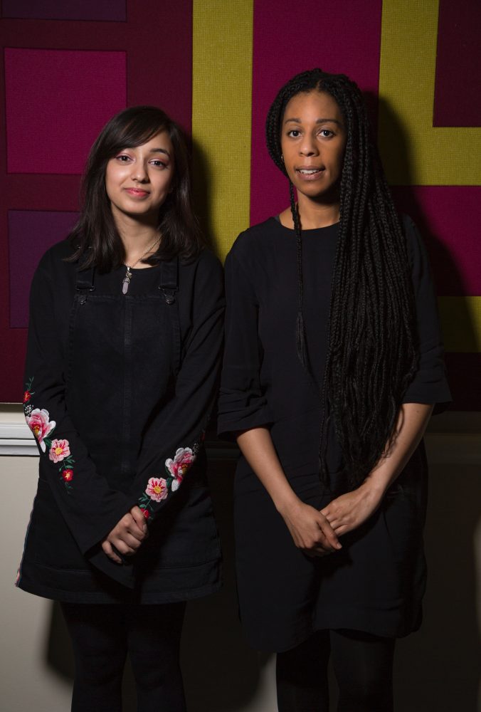 A young mentee and mentor stand together in front of a patterned green, pink, and purple wall. They are both clasping their hands in front of their bodies.