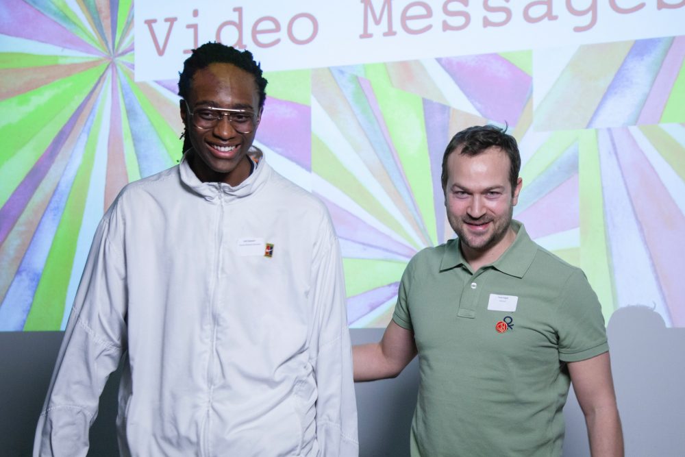 A young mentee stands on stage with his mentor against a bright green and pink projected background.