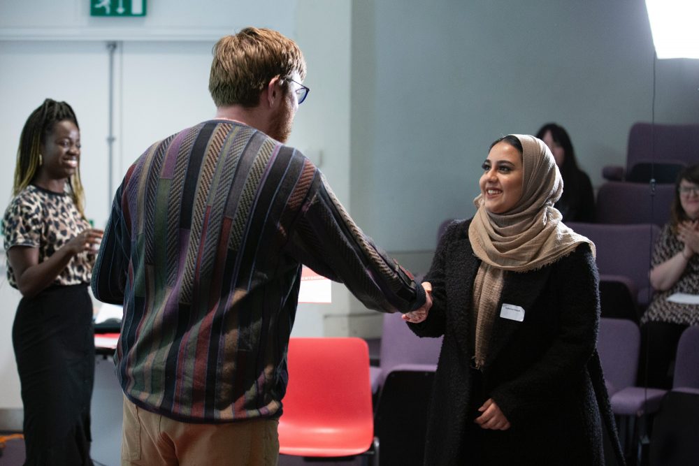 A young woman in a hijab smiles as she shakes hands with a man in a patterned jumper.