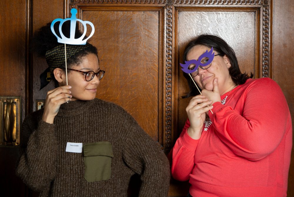 A young person and her mentor stand together. The mentee is holding a shiny, blue, paper crown on a stick above her head and looking at her mentor who is wearing a shiny, purple mask over her eyes.