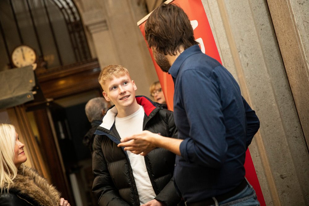 A young man in a white tshirt and black puffa jacket talking to two people.