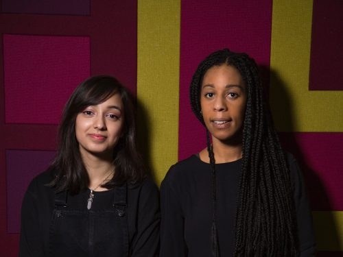 A young mentee and mentor stand together in front of a patterned green, pink, and purple wall. They are both clasping their hands in front of their bodies.