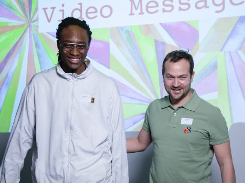 A young mentee stands on stage with his mentor against a bright green and pink projected background.