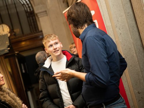 A young man in a white tshirt and black puffa jacket talking to two people.