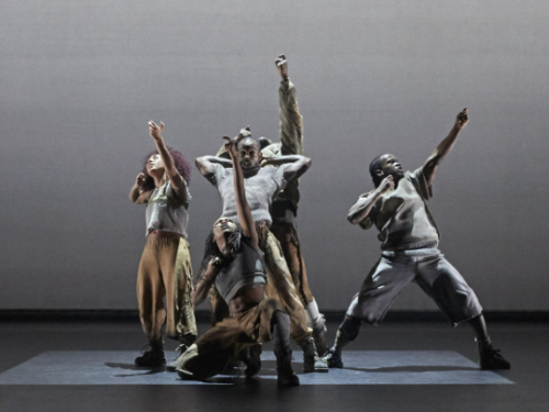 5 performers on stage wearing grey and beige trousers and t-shirts. They are standing in a square of light posing with arms in their air and look serious.