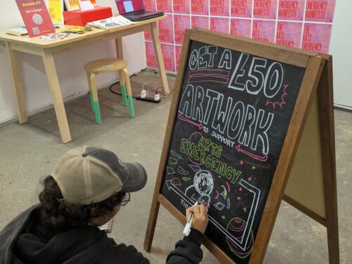Person kneeling on the floor decorating a large chalkboard sign at The Other Art Fair event