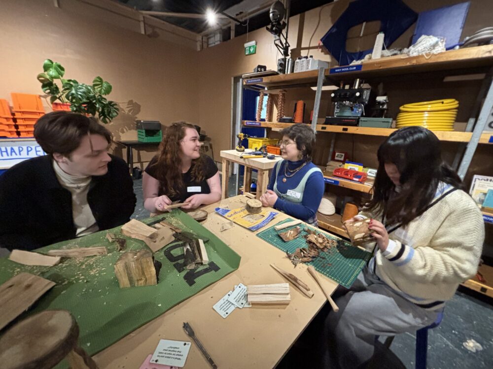 4 people sitting at a table whittling wood and chatting