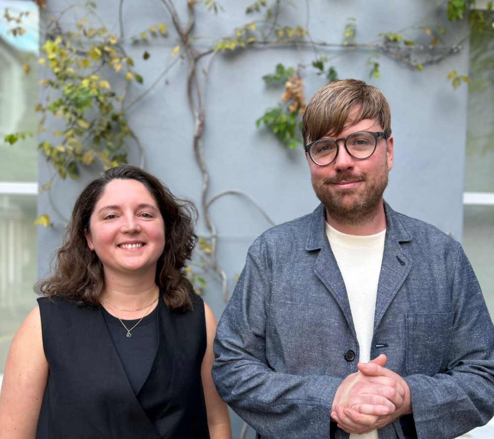 A female with shoulder length hair smiling and wearing a sleeveless black top standing next to a male with brown hair and a beard wearing glasses, a white t-shirt and a blue jacket