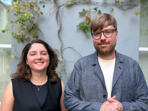 A female with shoulder length hair smiling and wearing a sleeveless black top standing next to a male with brown hair and a beard wearing glasses, a white t-shirt and a blue jacket