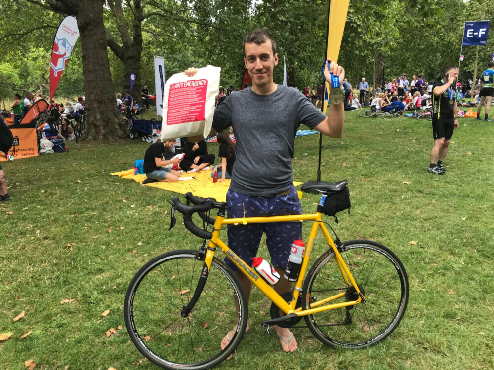 Person with their yellow bike leaning against them holds up their medal and an arts emergency packet in a busy park.