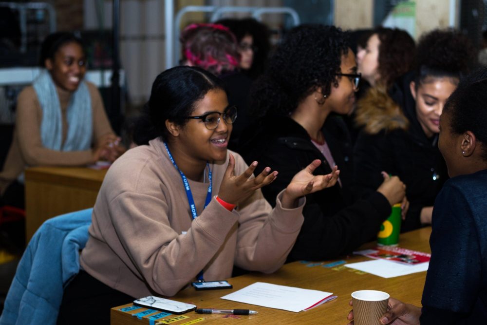 A young person gestures as she speaks to a woman sitting across the table from her, in a room with other similar pairs talking.