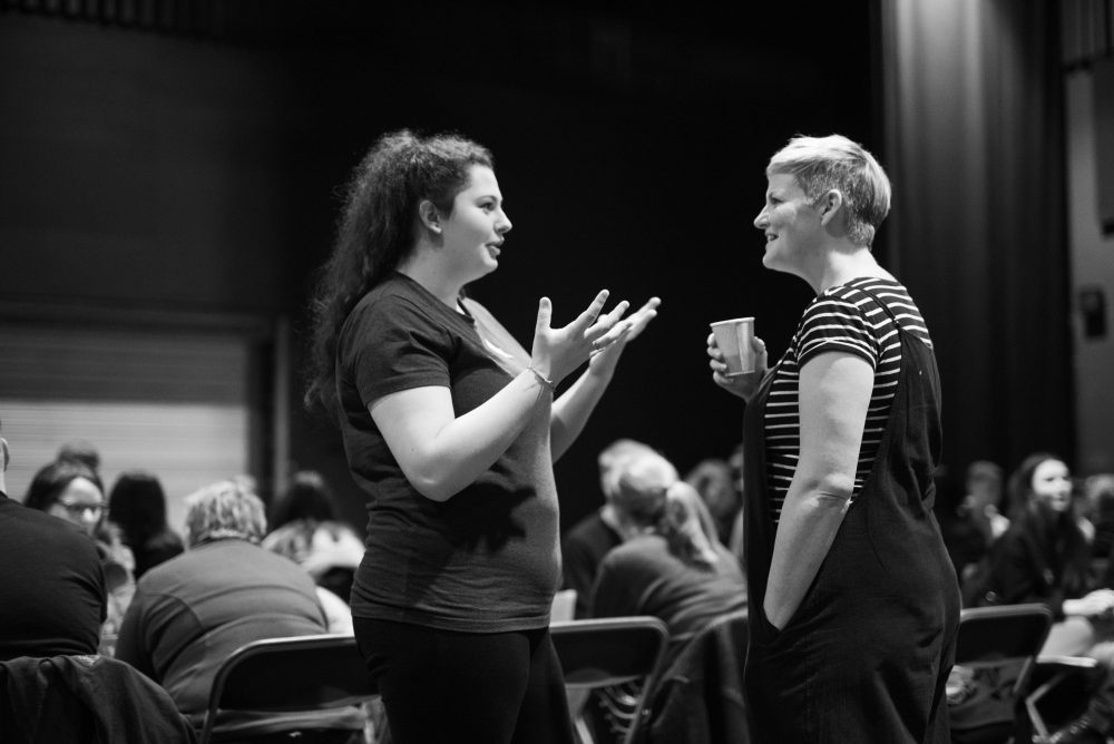 A young woman gestures with her hands as she speaks to a woman holding a paper cup.