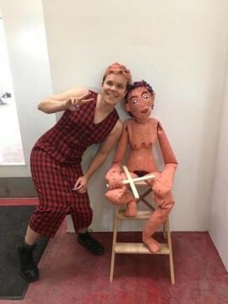 Susy  wearing a red checked dress stands and does a peace symbole next to a sculpture of a person sat on a chair.