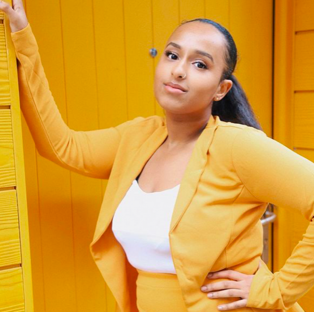 Young Community member Abrehet leans in a sunny, bright yellow doorway, wearing a sunny, bright yellow suit. She has her hair pulled back and is smiling slightly.
