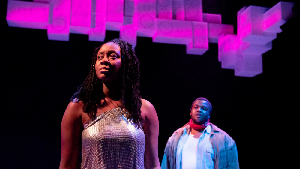 A Black woman and Black man look into the distance on a black set with speakers above them