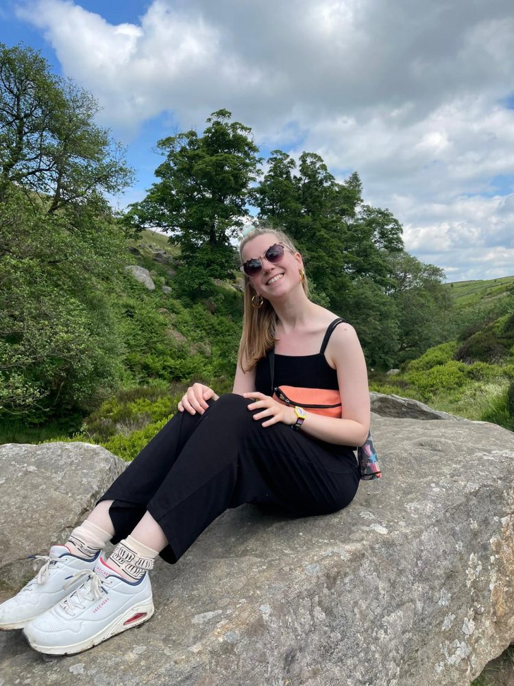A picture of writer Bryony Taylor. She is a woman with white skin and mid-length blonde hair. She is sitting ona rock in a natural landscape wearing trainers and a black outfit, holdin an orange bag and wearing sunglasses. She is smiling.