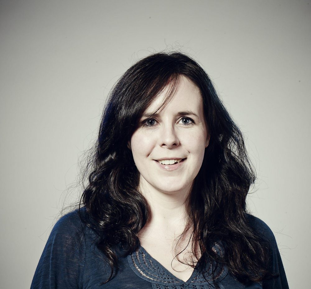 A portrait of writer Helen O'Hara. She is smiling and has pale white skin and mid-length curled black hair and is wearing a blue top with lace decoration. She is standing against a grey background and the corners of the image have a shadow vignette darkening them.