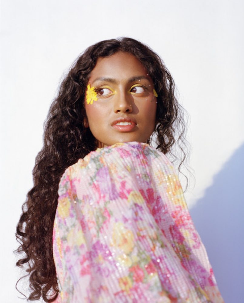 A brown girl with long curly hair and a floaty pink floral top looking over her shoulder against a white wall in bright sunlight. She has a flower on her face and sunny makeup.