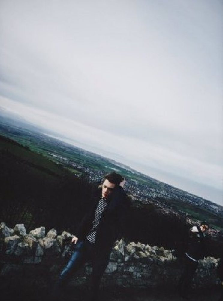 A young man stands with a stone wall behind him and rolling hills.