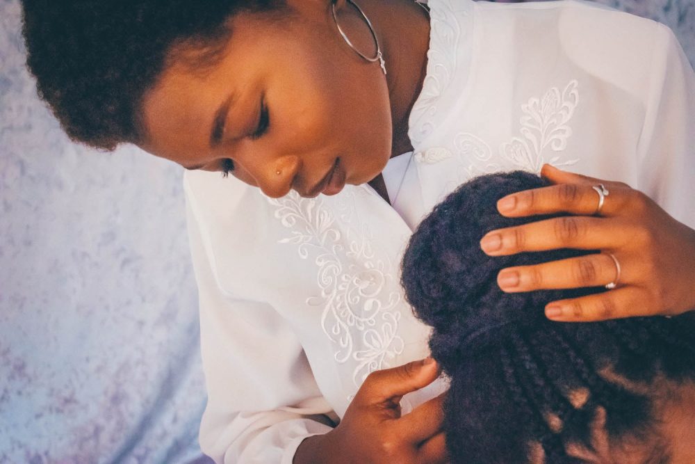 A young Black woman wearing a white top and hoop earrings leans over another Black woman and places her hands gently on her hair.