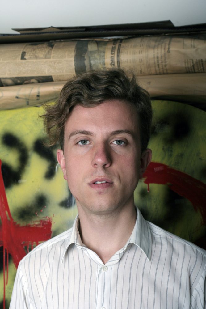 A head and shoulders view of a man wearing a white shirt, standing in front of a spray painted wall