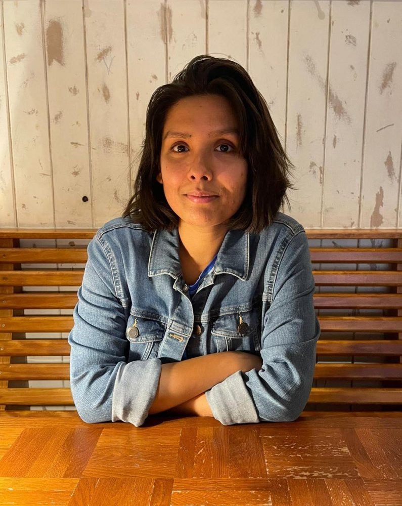 A picture of writer Shyama Laxman who is a woman with brown skin and black hair cut in a bob. She is wearing a denim jacket and sitting at a wooden table with her crossed arms leaning on the surface. She is smiling at the camera.