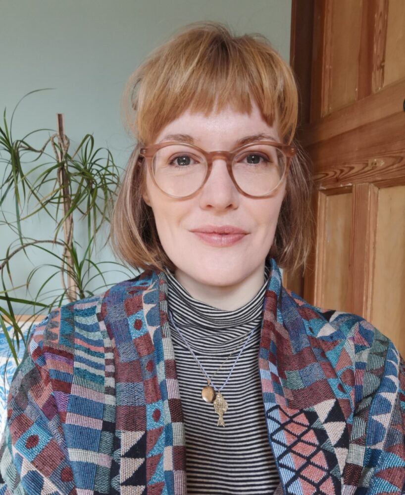 A woman with blond shoulder length hair and a fringe. She is wearing large round glasses, a patterned cardigan and a grey polo neck.