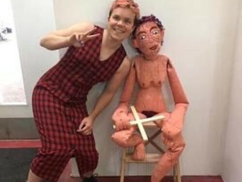 Susy  wearing a red checked dress stands and does a peace symbole next to a sculpture of a person sat on a chair.