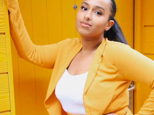 Young Community member Abrehet leans in a sunny, bright yellow doorway, wearing a sunny, bright yellow suit. She has her hair pulled back and is smiling slightly.