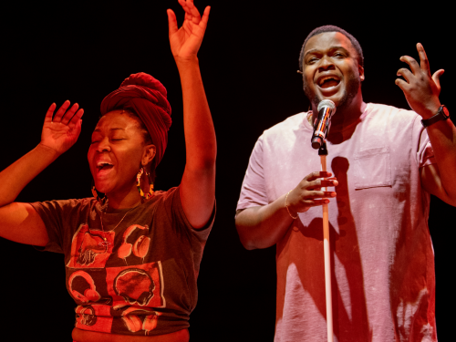 A Black woman raises her arms and sings and a Black man sings with a microphone