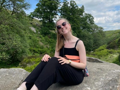 A picture of writer Bryony Taylor. She is a woman with white skin and mid-length blonde hair. She is sitting ona rock in a natural landscape wearing trainers and a black outfit, holdin an orange bag and wearing sunglasses. She is smiling.