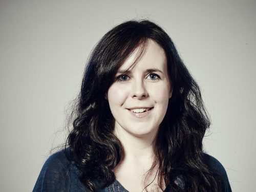 A portrait of writer Helen O'Hara. She is smiling and has pale white skin and mid-length curled black hair and is wearing a blue top with lace decoration. She is standing against a grey background and the corners of the image have a shadow vignette darkening them.