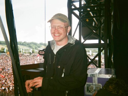 A white male wearing a beige baseball cap and black jacket, smiling. He is backstage at a festival.