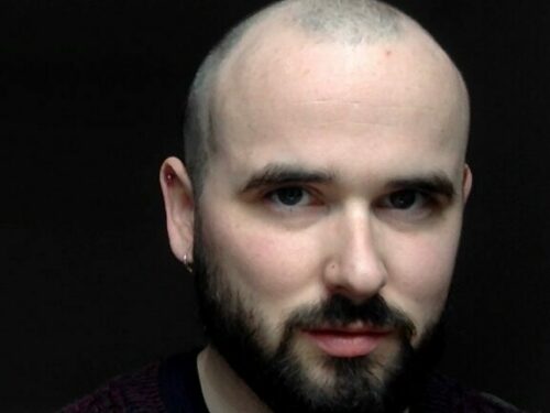 A portrait of author Michael Magee. Against a deep black background the author's head and shoulders. Michael is wearing a black t-shirt, he has white skin, a shaved head and a black beard and moustache. He is smiling slightly with his face turned a few degree towards the right hand side of the frame.