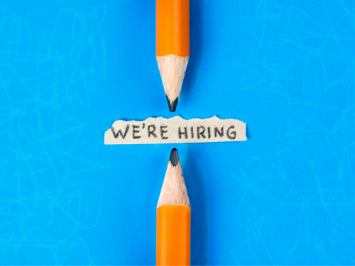 A scrap of paper reads 'we're hiring' and two pencils point to it from the top and the bottom. The background is blue.