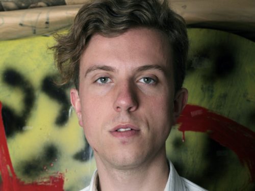 A head and shoulders view of a man wearing a white shirt, standing in front of a spray painted wall