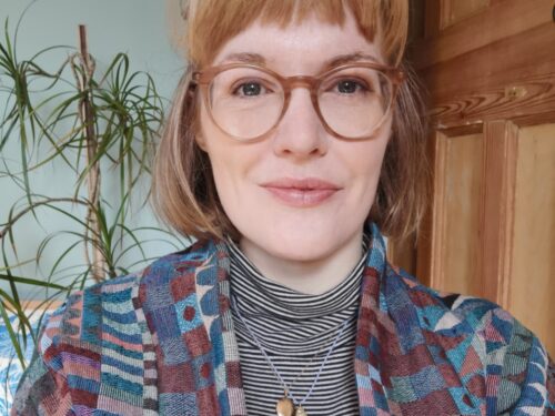 A woman with blond shoulder length hair and a fringe. She is wearing large round glasses, a patterned cardigan and a grey polo neck.