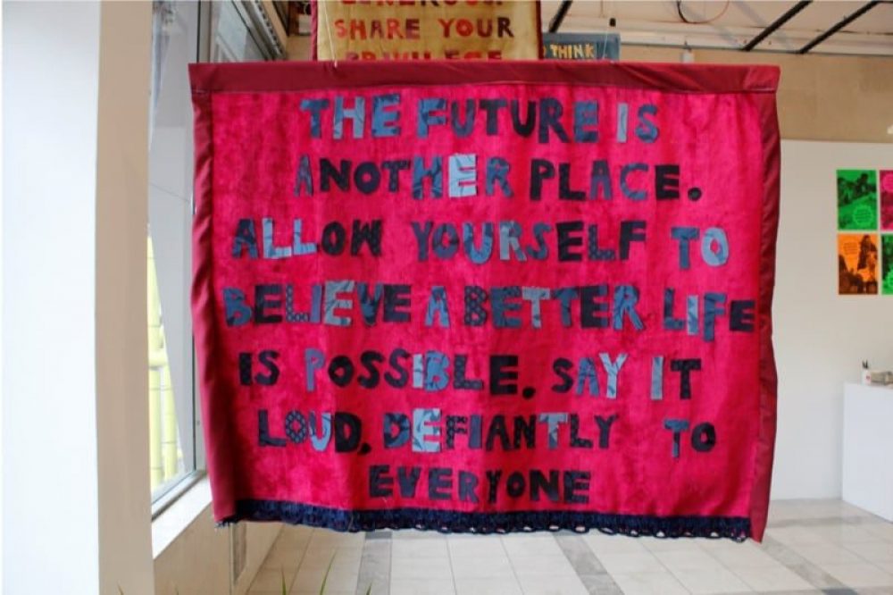 A red banner hangs on display at an exhibition. In blue applique letters the banner reads ‘The Future is another place. Allow yourself to believe a better life is possible. Say it loud defiantly to everyone.’