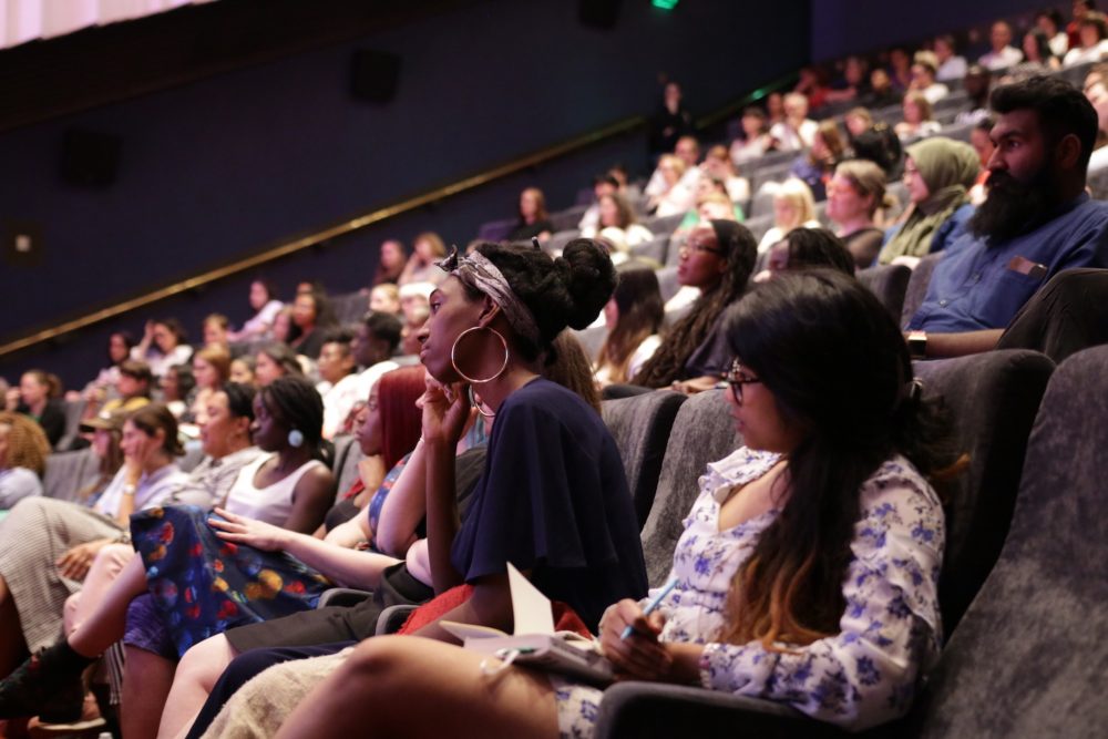 An audience of young people in theatre seats look towards the stage, some take notes.